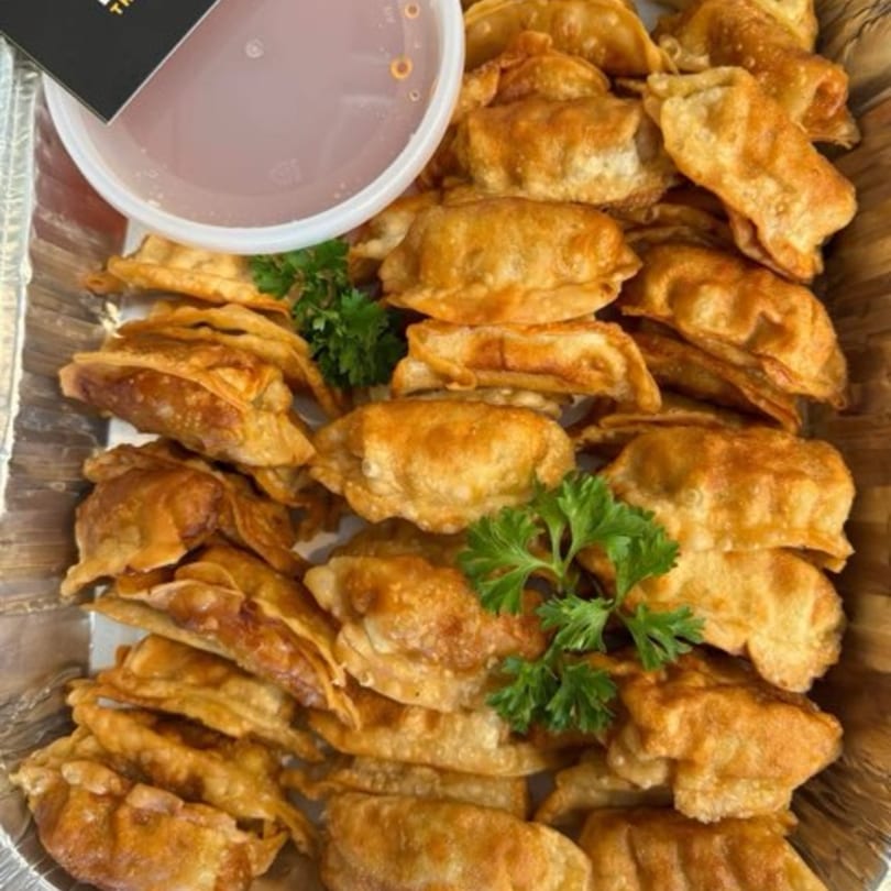 Crispy fried Thai dumplings with fresh parsley in a foil tray, with a side of dipping sauce.