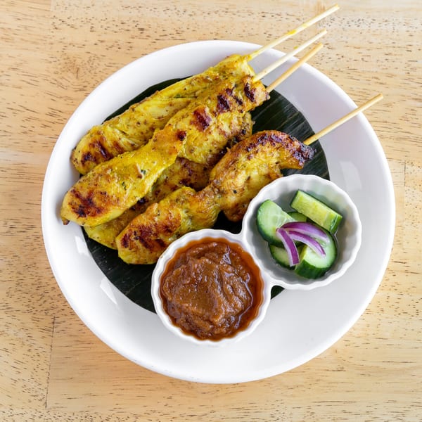 Grilled chicken satay skewers with peanut sauce and cucumber salad on a white plate.
