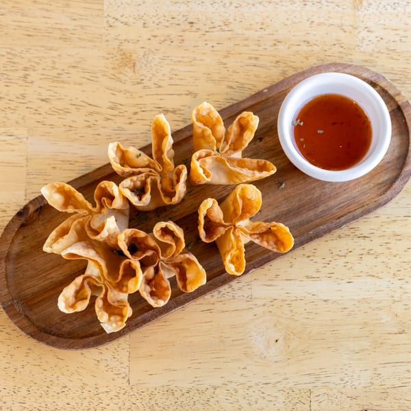 Crispy golden crab rangoons, shaped like flowers, with a side of sweet chili sauce on a wooden tray.