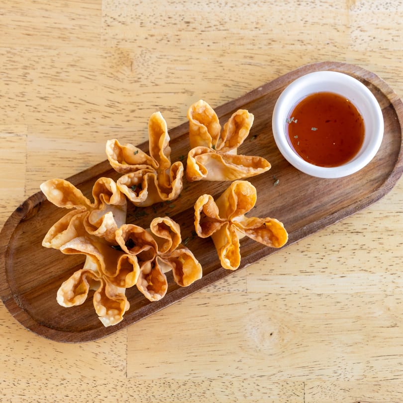 Crispy golden crab rangoons, shaped like flowers, with a side of sweet chili sauce on a wooden tray.
